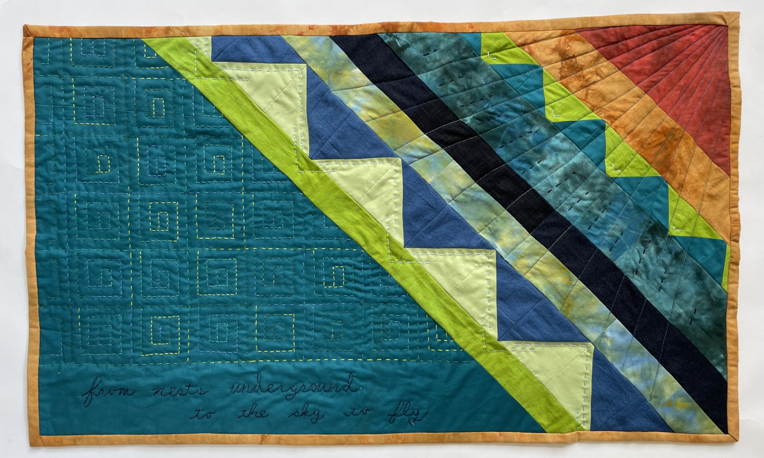 pieced quilt with bird tail at upper right like a sunburst and text that says from nests in the ground to the sky to fly