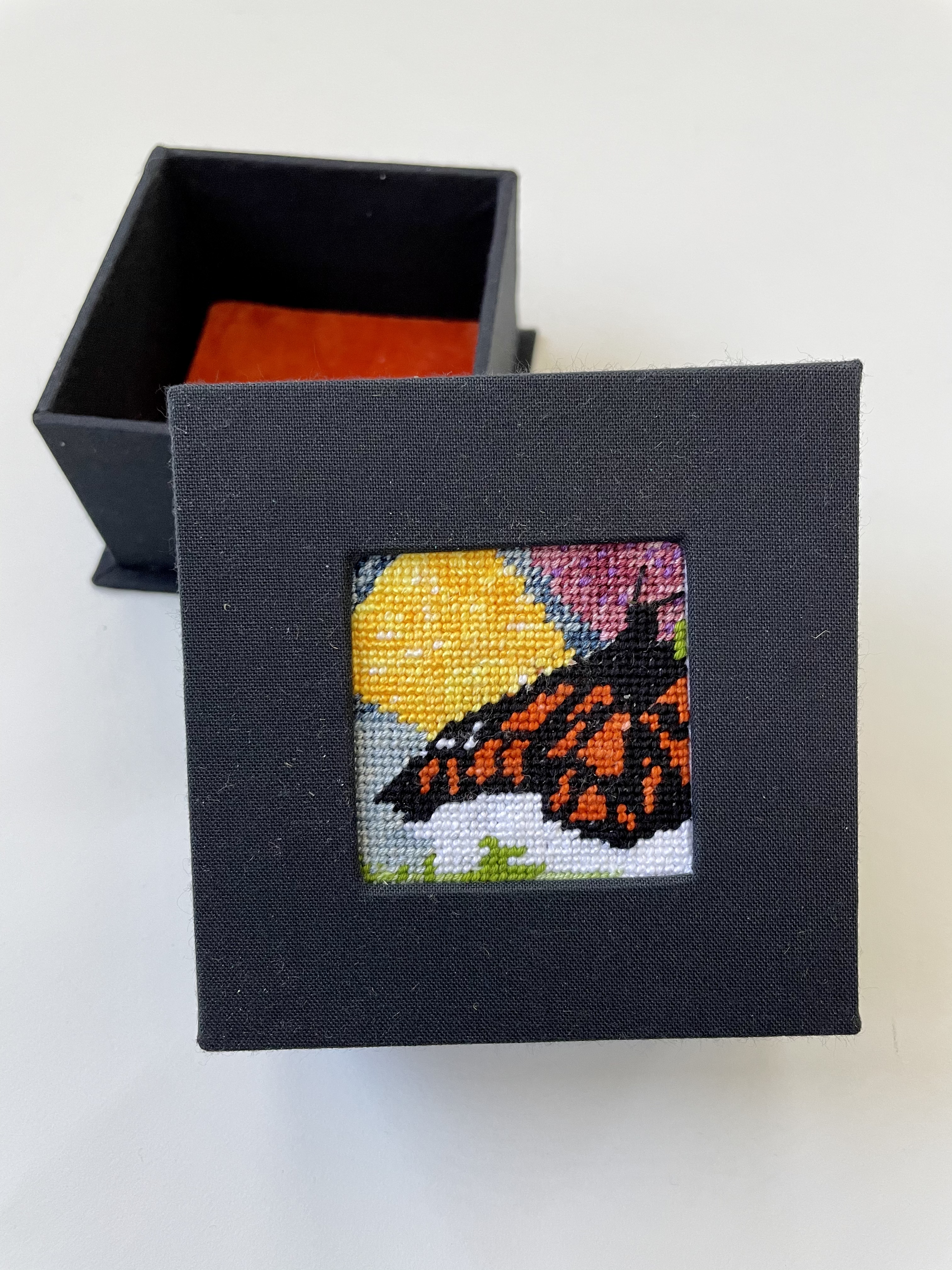 a box with an inset needlepoint of a monarch butterfly against abstract flower and sun and grass